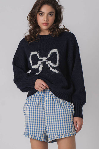 A woman wearing a navy blue crewneck sweater with a bow graphic and white checkered shorts.