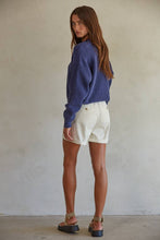 Load image into Gallery viewer, A woman is wearing a blue button down cardigan with brown buttons over a white top, paired with beige shorts. She is posing against a neutral background.
